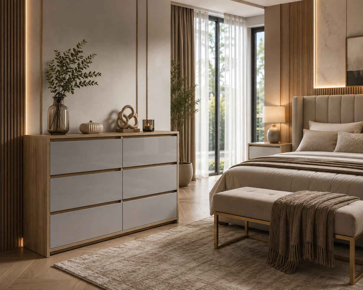 A modern bedroom scene featuring a light wood and white dresser with gold-toned handles, styled with decorative vases and a plant. The room includes a bed with neutral bedding, a matching upholstered bench, a rug, and warm ambient lighting.